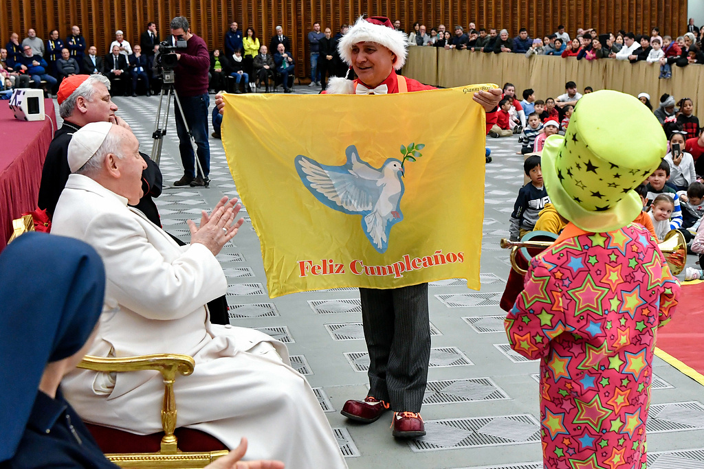 Papst Franziskus während einer Audienz mit betreuten Kindern von der Krankenstation Santa Marta an seinem 87. Geburtstag am 17. Dezember 2023 im Vatikan. Ein Mann zeigt ihm eine Fahne mit der Aufschrift "Feliz Cumpleanos" (dt. Herzlichen Glückwunsch zum Geburtstag). (Foto: KNA)